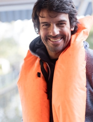 A man with a joyful expression is wearing an orange life jacket over a dark hoodie. The background is blurred, suggesting an outdoor setting.