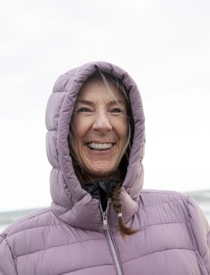Smiling woman wearing a lavender puffer jacket with the hood up stands outdoors, with a cloudy sky in the background.