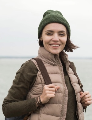 A person smiles warmly while standing outdoors near the water. They wear a green beanie, a brown puffer vest, and carry a backpack. The sky is overcast, and the background features a body of water.