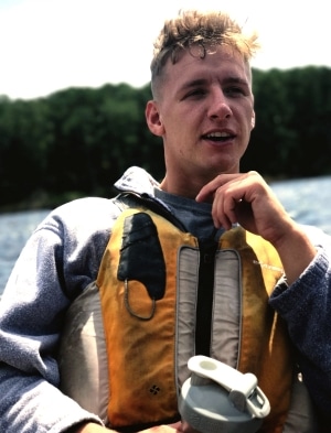 Man wearing a life jacket sits on a boat with water and trees in the background. He's holding a water bottle and looking towards the side. The scene is bright and outdoorsy.