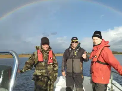 Three people in life jackets stand on a small boat in calm waters, smiling under a clear sky. A colorful rainbow arcs overhead, adding to the scenic backdrop.