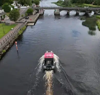 A pink amphibious vehicle drives across a river in a town with a stone bridge in the background. Lush greenery and buildings line the riverbanks under a cloudy sky.