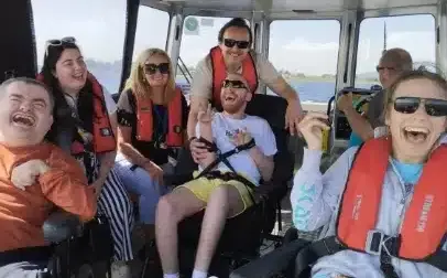 A group of smiling people wearing life jackets are on a boat. The sun is shining, and they seem to be enjoying their time together on the water. Two individuals are in wheelchairs, and everyone looks happy.
