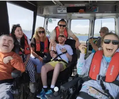 A group of smiling people wearing life jackets are on a boat. The sun is shining, and they seem to be enjoying their time together on the water. Two individuals are in wheelchairs, and everyone looks happy.