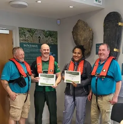 Four people wearing life jackets smile and hold certificates indoors. Two large historical artifacts are displayed on the wall behind them. The group appears to be celebrating an achievement.