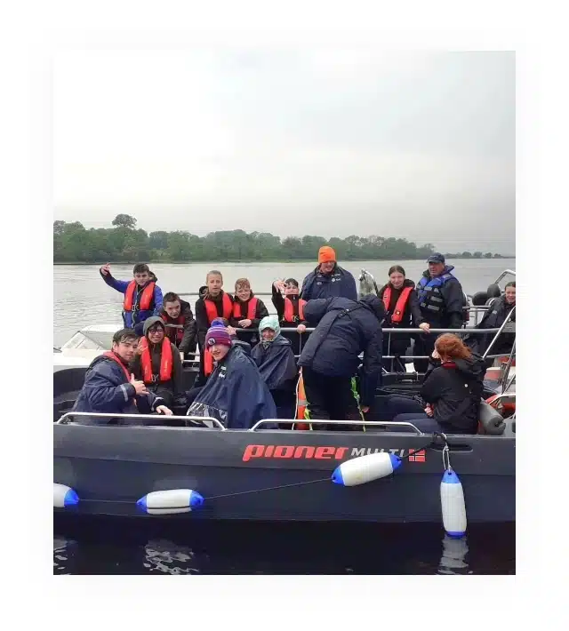 A group of people wearing life jackets are seated in a motorboat on a calm body of water. The boat is labeled "Pioneer Multi." Overcast skies and distant trees are visible in the background. Some people are standing and holding onto the boat's rails.