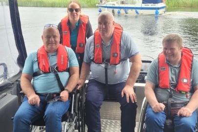 Four people wearing red life jackets, two in wheelchairs, on a boat with water and reeds in the background. A boat is visible on the water behind them.