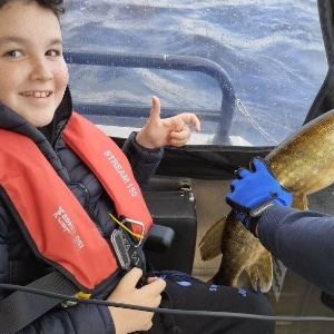 A person wearing a red life jacket is sitting in a boat, smiling and giving a thumbs-up. Another person wearing blue gloves is holding a large fish inside the boat, against a backdrop of water visible through a clear plastic cover.