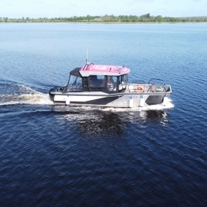 A small motorboat with a canopy and a pink roof cruises across a calm, expansive blue lake. Green trees and a clear sky are visible in the distant background.