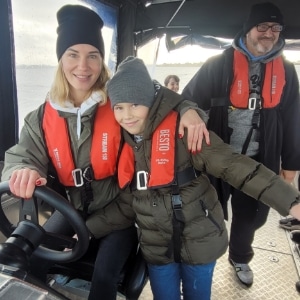 A woman and a child wearing life jackets are on a boat. The woman is seated at the helm with her hand on the wheel, while the child stands close, smiling. A man stands behind them, also wearing a life jacket. They all appear to be dressed for cold weather.
