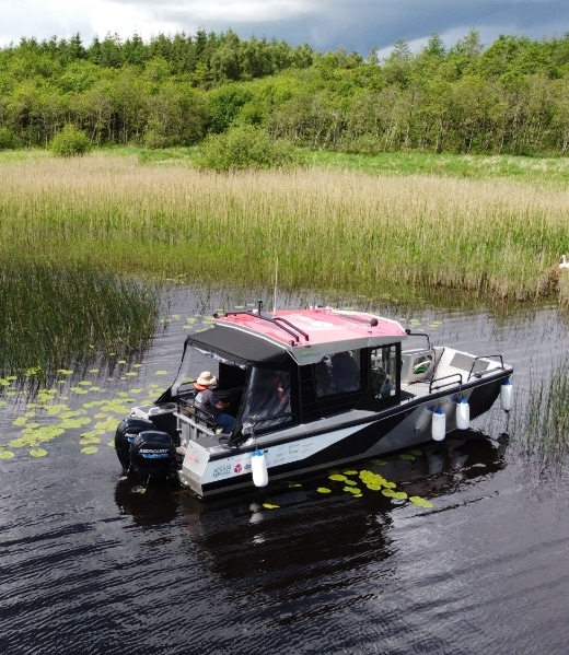 A small motorboat with a red canopy floats on a calm lake surrounded by tall reeds and lily pads. A person in a hat is seated inside, steering the boat. A lush, green forest is visible in the background under a cloudy sky.
