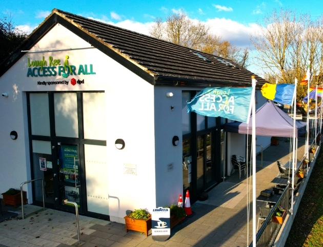 A small, white building with a sign reading "Access for All" and colorful flags lining a walkway. The sky is clear with a few clouds, and trees are visible in the background. The building appears welcoming and accessible.