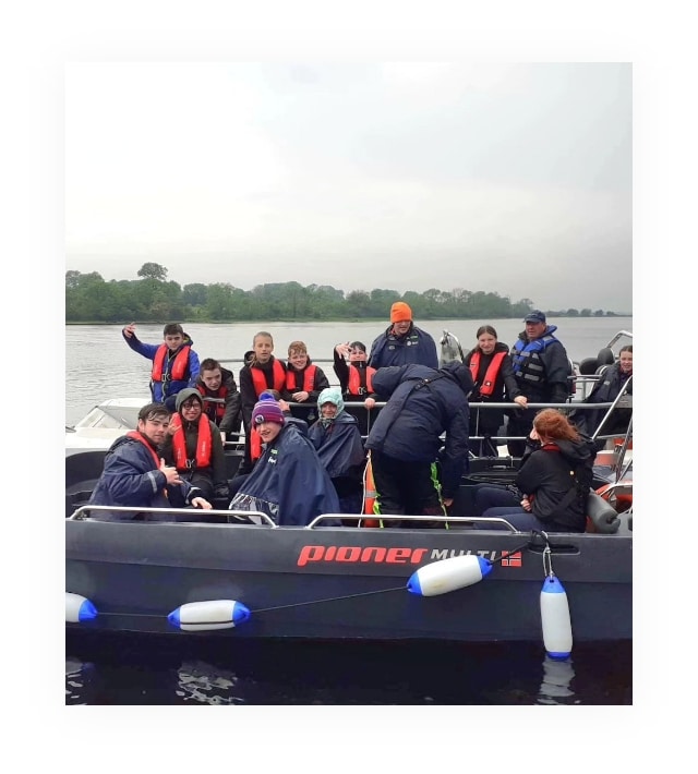 A group of people wearing life jackets sit in a small boat on a body of water. Some are smiling and waving at the camera. The boat has several passengers and is equipped with safety floats. The background shows a cloudy sky and distant trees.