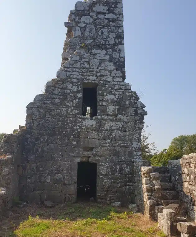 Old stone building ruins with a tall, narrow chimney and openings for doors and windows. The structure is weathered and partially overgrown with moss. Bright sky in the background, surrounded by sparse greenery.