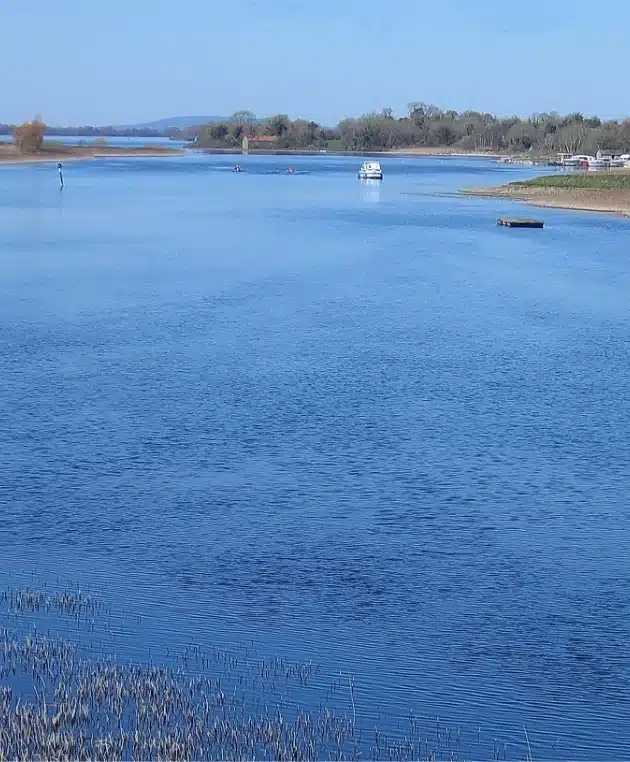 A tranquil blue river with a boat in the distance, surrounded by grassy banks and trees. The sky is clear and sunny, reflecting on the water's surface.