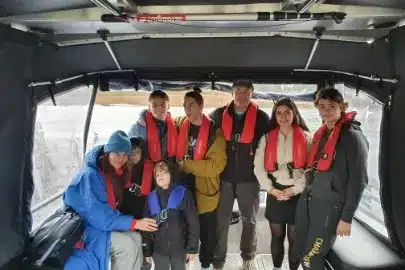 A group of seven people, including young children and adults, are on a boat wearing life jackets. They are smiling and dressed in warm clothing, with a snowy landscape visible in the background.