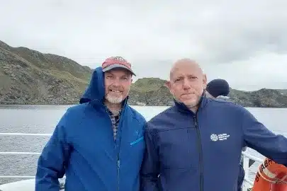 Two men stand on a boat in front of rocky hills by the water. The man on the left wears a red cap and blue jacket, while the man on the right wears a blue jacket with a white logo. The sky is cloudy, and a life preserver is visible nearby.