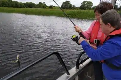 Two people in life jackets are on a boat, engaged in fishing. One person is helping the other reel in a fish from the water. The scene is surrounded by greenery on the far shore.