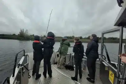 A group of people wearing jackets and life jackets are fishing from a boat on a cloudy day. They are standing on the deck, holding fishing rods, with a view of the water and distant shore in the background.