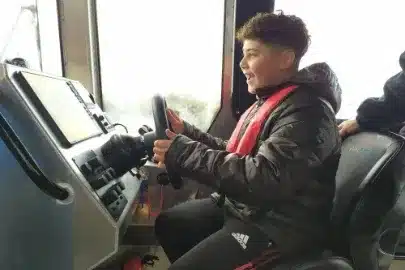 A young person with curly hair is smiling while sitting in the captain's seat of a boat, holding the steering wheel. They are wearing a dark jacket and a red life jacket. The interior of the boat is visible with windows showing the outside.