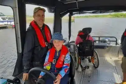 A man and a child, both wearing life jackets, smile on a boat. The man stands behind the steering wheel, and the child sits beside him wearing a blue hat. Another person in a wheelchair is visible in the background. The boat is on a calm body of water.