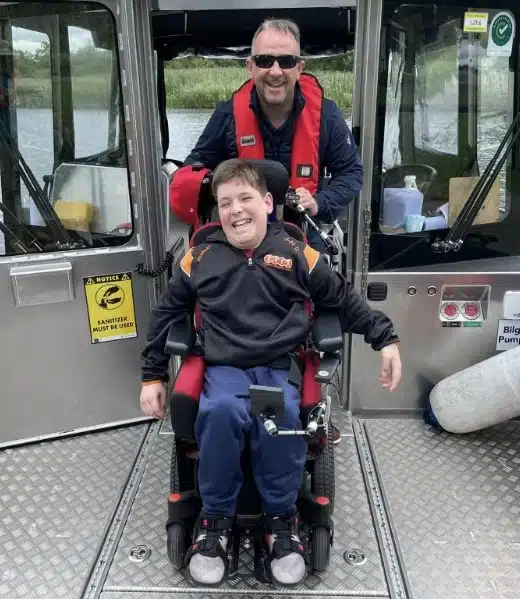 A smiling boy in a wheelchair is in front of a man wearing a red life vest. They are on a metal boat, with water and greenery visible in the background. Various signs and a sanitizer bottle are visible on the boat.