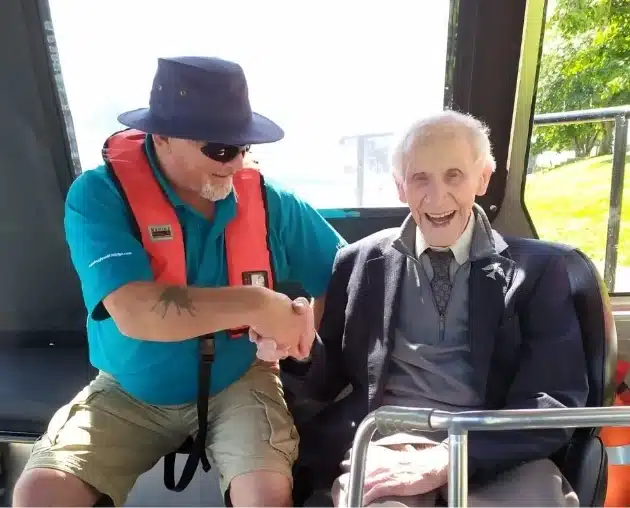 A man wearing a blue hat and life vest smiles and shakes hands with an elderly man in a blazer on a boat. The elderly man, also smiling, is seated on the right. Sunlight illuminates the scene through the window.