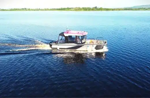 A small motorboat with a pink roof travels on a vast, calm lake. The water's surface is slightly rippled by the boat's movement. Green trees and a clear sky are visible in the background.
