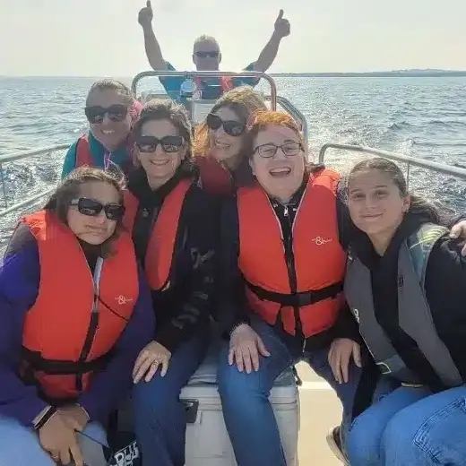 A group of five people wearing life jackets are smiling and sitting in a small boat on a sunny day. A man in the background gives a thumbs-up while steering the boat. The water and sky are visible in the background.