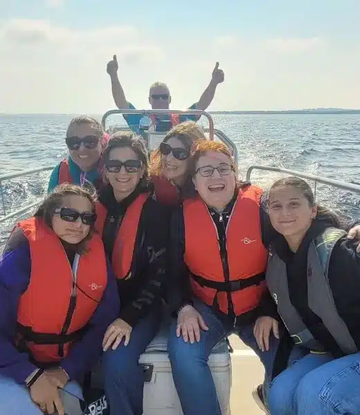 A group of five people wearing life jackets are smiling and sitting in a small boat on a sunny day. A man in the background gives a thumbs-up while steering the boat. The water and sky are visible in the background.