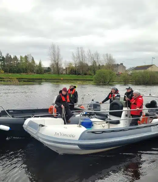 A group of people in life jackets are on two motorboats on a calm river. One boat is named "Invincible". Trees and a few buildings are visible in the background under a cloudy sky.