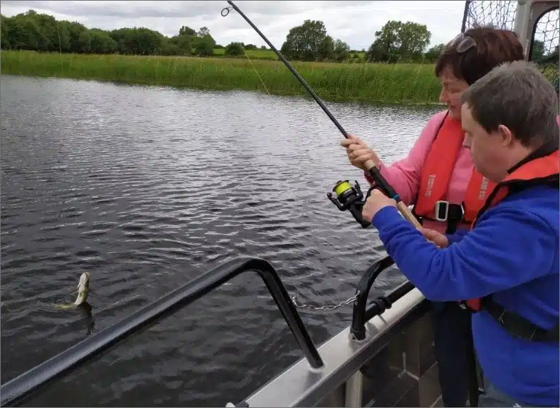 Two people are on a boat fishing. One is holding a fishing rod with a fish on the line, wearing red life vests. Water and reeds are visible in the background under a cloudy sky.