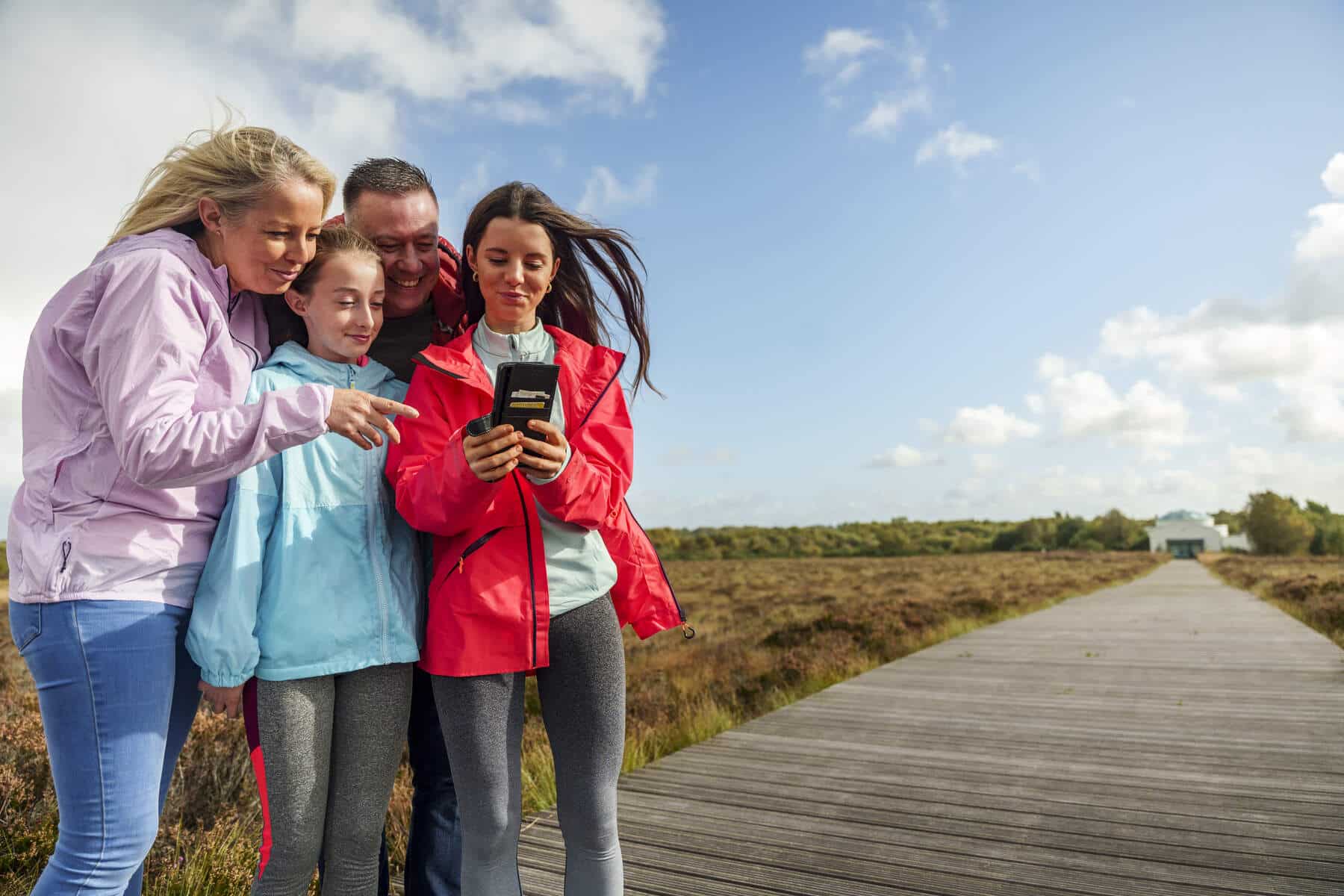 A family of four stands on a boardwalk in a natural landscape, smiling and looking at a handheld device together under a blue sky—enjoying Accessible Trip Planning for Lough Ree.