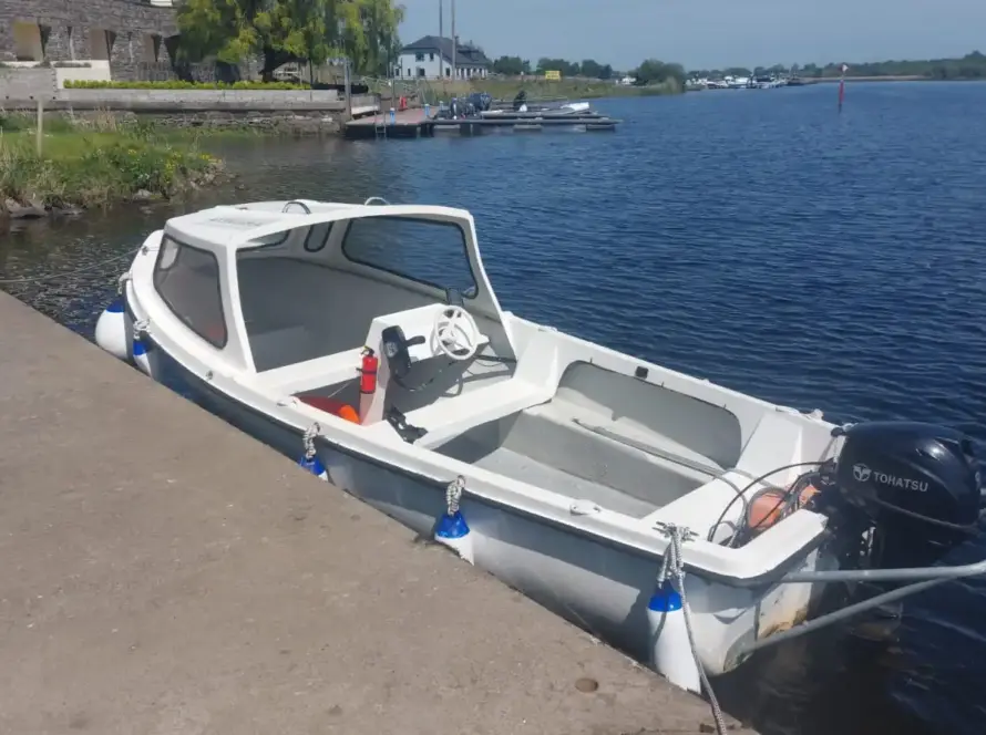 A small white motorboat with a canopy, part of Accessible Lake Boat Hire Lough Ree, is moored to a concrete dock on a calm river, with buildings and other boats visible in the background under a clear sky.