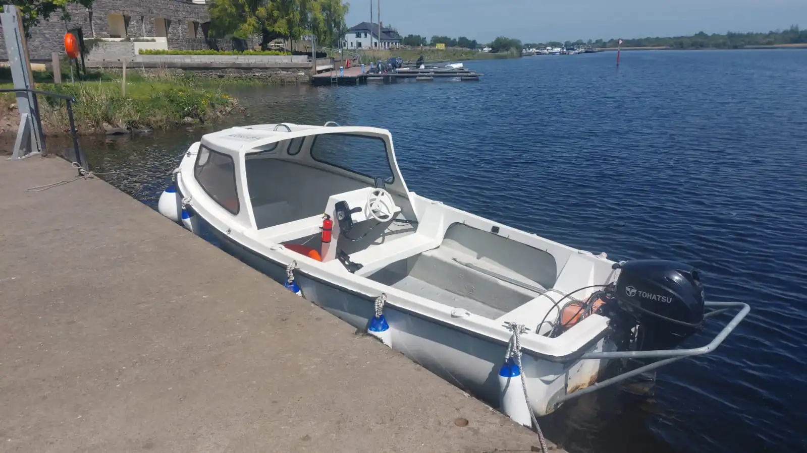A small white motorboat with a canopy, part of Accessible Lake Boat Hire Lough Ree, is moored to a concrete dock on a calm river, with buildings and other boats visible in the background under a clear sky.