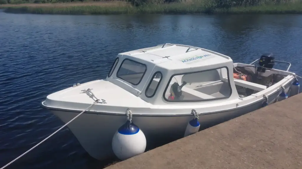 A small white motorboat with a closed cabin is moored to a concrete dock on a calm river, blue fenders hanging on the side and reeds in the background—an ideal option for Accessible Lake Boat Hire on Lough Ree.