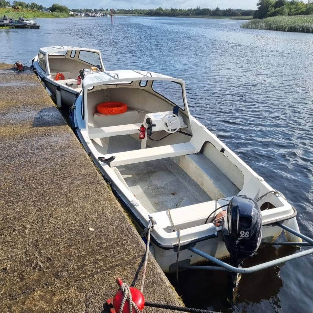 Two small white motorboats, part of Accessible Boat Trips Roscommon, are tied to a concrete jetty on a calm river. Life rings and fire extinguishers are visible inside the boats, with lush green vegetation lining the opposite riverbank.