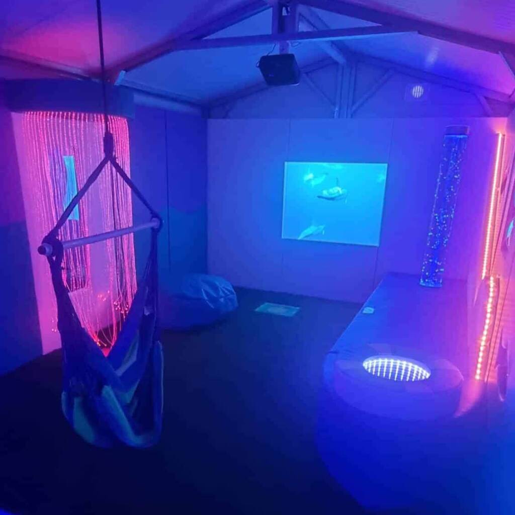 A sensory room with purple and blue lighting features a hanging chair, a bean bag, a bubble tube, fiber optic lights, and a wall projection of underwater scenes, creating a calming, immersive environment.