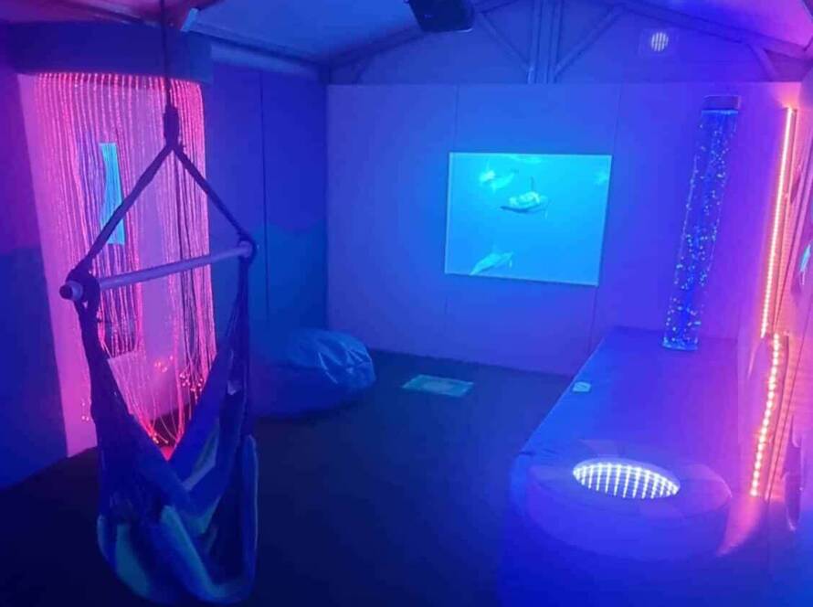 A sensory room with purple and blue lighting features a hanging chair, a bean bag, a bubble tube, fiber optic lights, and a wall projection of underwater scenes, creating a calming, immersive environment.