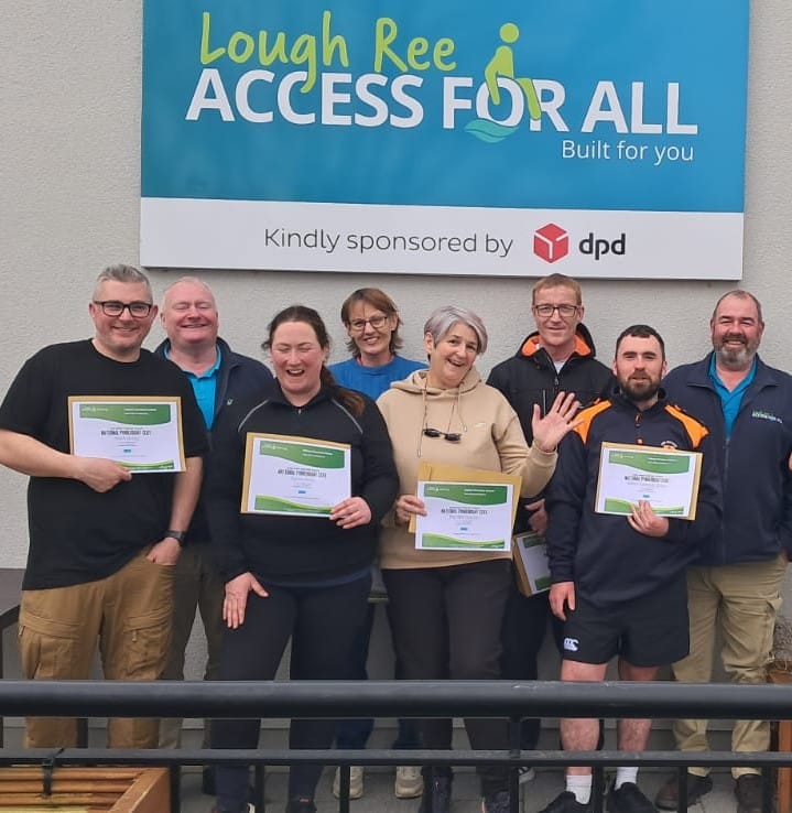 A group of eight smiling people holding certificates pose in front of a sign that reads "Lough Ree Access For All, Built for you, Kindly sponsored by dpd," celebrating accessible corporate group activities at Lough Ree.